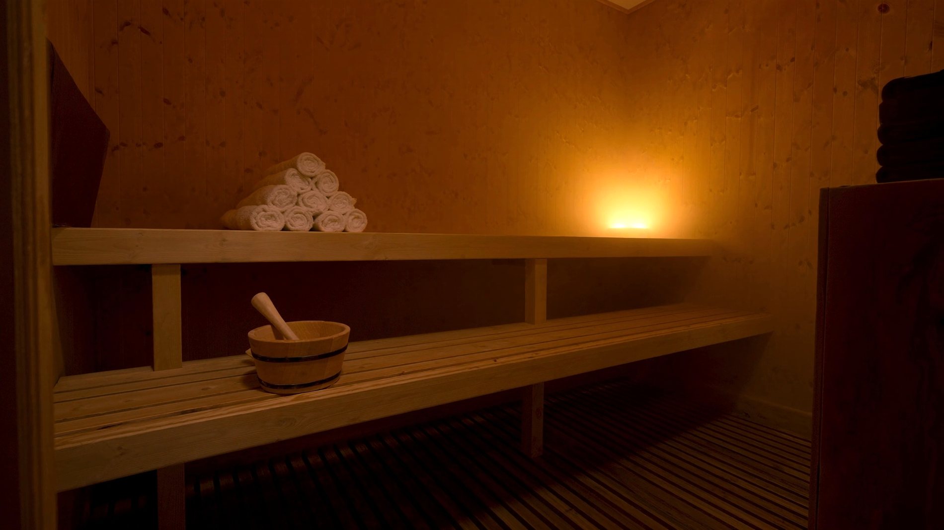 The cedar room in low light: two tiers of plank bench, a wooden bucket with a ladle sitting on the lower bench, and a stack of rolled white towels on the upper one.