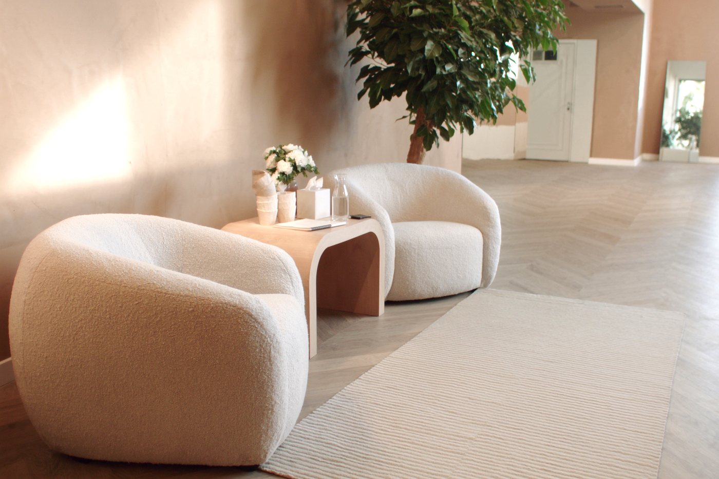 The front room: two low chairs in oatmeal fabric, a round oak side table with a glass water carafe and stacked cups, and a large potted plant against a warm plaster wall.
