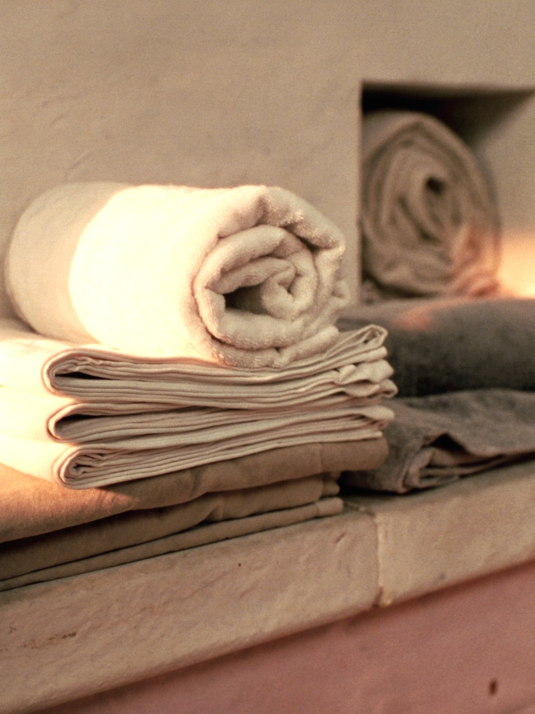 Rolled cream towels and folded linen sheets stacked on a pale stone shelf in warm side light.