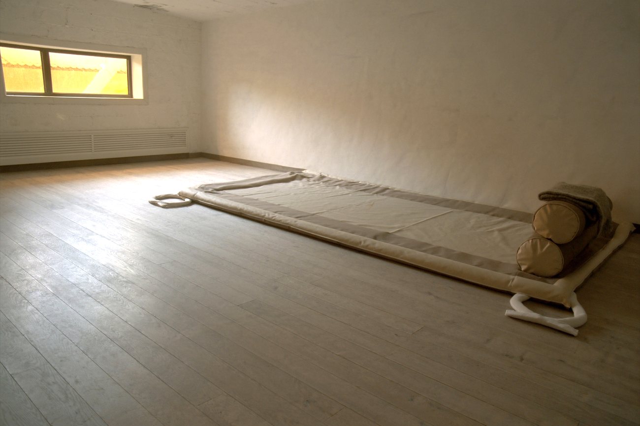 The mat room: a wide padded floor mat laid out on pale board floors, two firm bolsters and a folded wool blanket stacked at one end, light from a low window.