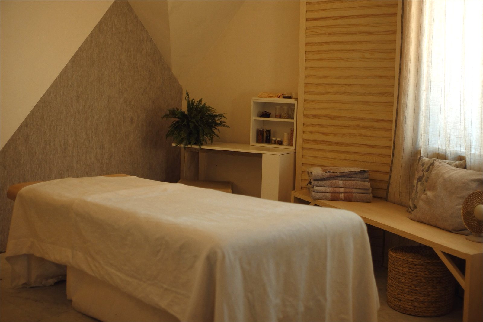A treatment room in the late morning: a low table made up in cream linen, a stack of folded sheets on a pale oak bench, and a slatted oak wall behind it.