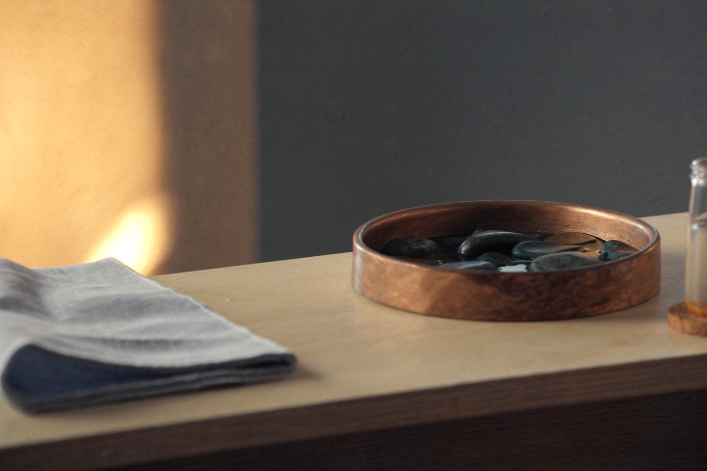 Smooth dark river stones warming in a shallow copper bowl of water on an oak counter, a folded linen cloth beside it.